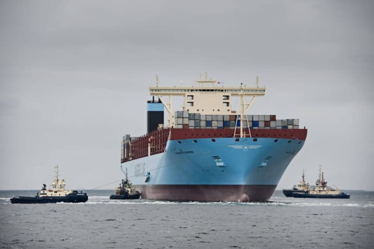 A head-on view of a massive MAERSK cargo ship being guided through the water by four tugboats