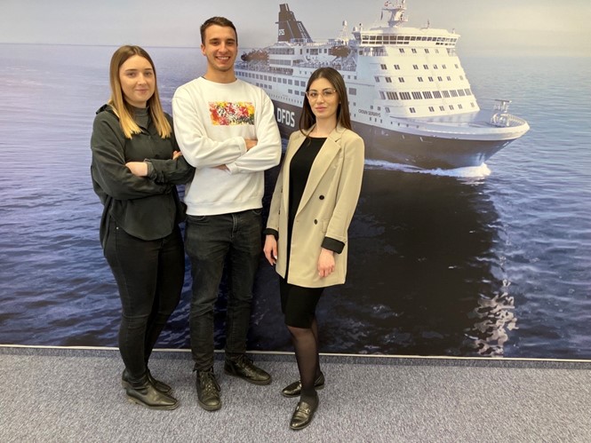 A picture of three DFDS employees standing together in front of a large image of a DFDS passenger ferry