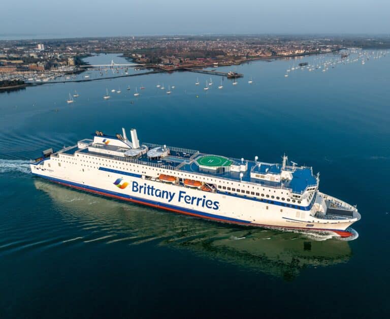 A wide shot of a Brittany Ferries vessel sailing away from land, with the coastline still visible in the background