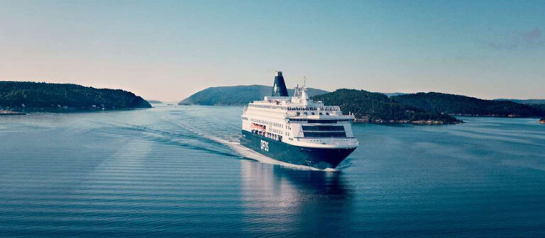 A DFDS passenger ship sails through a narrow waterway surrounded by forested hills under a clear sky