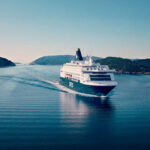 A DFDS passenger ship sails through a narrow waterway surrounded by forested hills under a clear sky