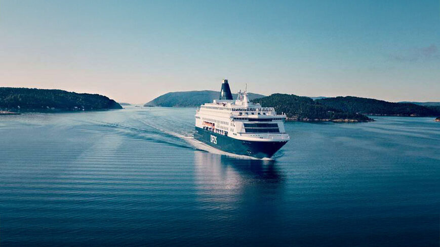A DFDS passenger ship sails through a narrow waterway surrounded by forested hills under a clear sky