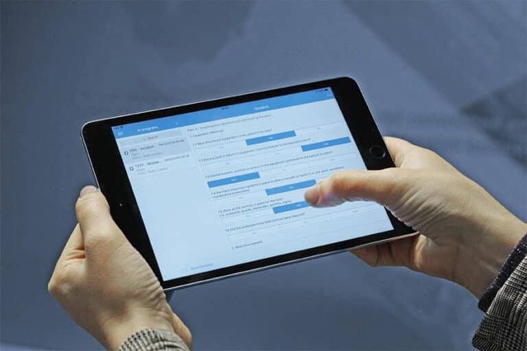 A tablet displaying the SERTICA app on its screen is being held by a pair of hands, with the interface clearly visible