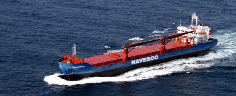A sizable blue and red cargo vessel sails across the ocean, leaving a wake in its path