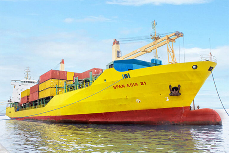 A yellow and red tanker vessel with stacks of colorful containers in various shades of blue, red, and green, neatly stacked in rows, is docking at a port