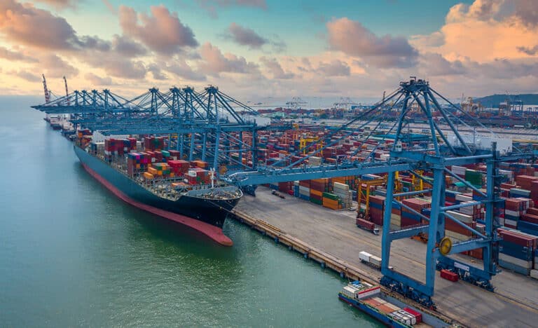 A large cargo vessel anchored at a harbor, with numerous containers stacked in neat rows on the dock