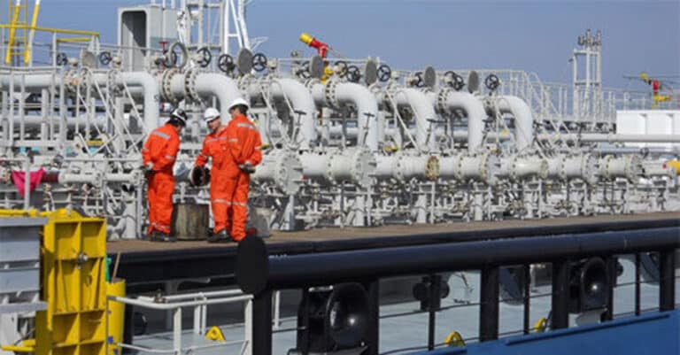 Three men in orange uniforms work on a ship, handling pipes and equipment in a maritime environment