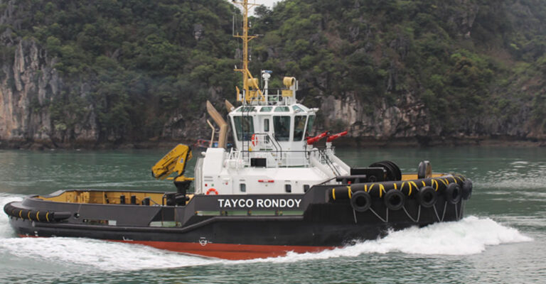 A side view of a tugboat sailing close to the land, with mountains in the background